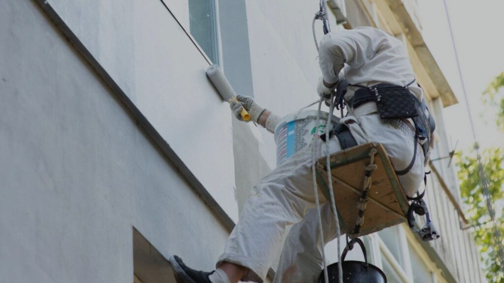 Profissional de pintura em cadeira suspensa limpando e recompondo a fachada de edifício com rolo de pintura e balde de tinta.