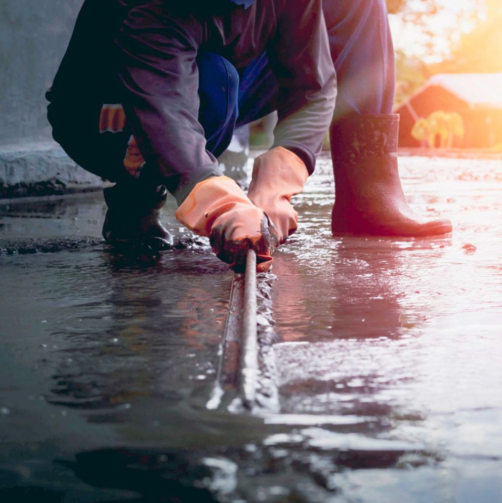 Profissional agachado aplicando manta líquida de impermeabilização em juntas de piso, usando luvas de borracha e botas de segurança.
