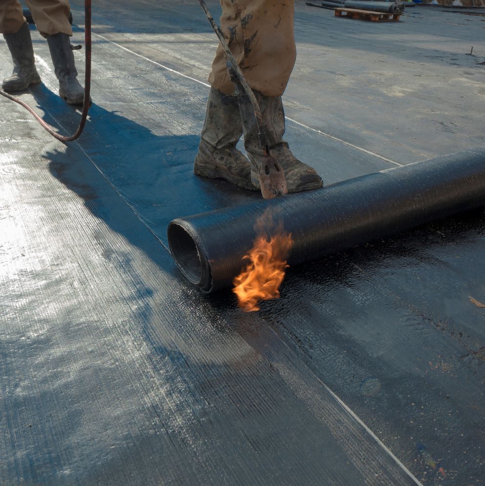 Operários aplicando manta asfáltica com maçarico para impermeabilização da fundação de edifício.