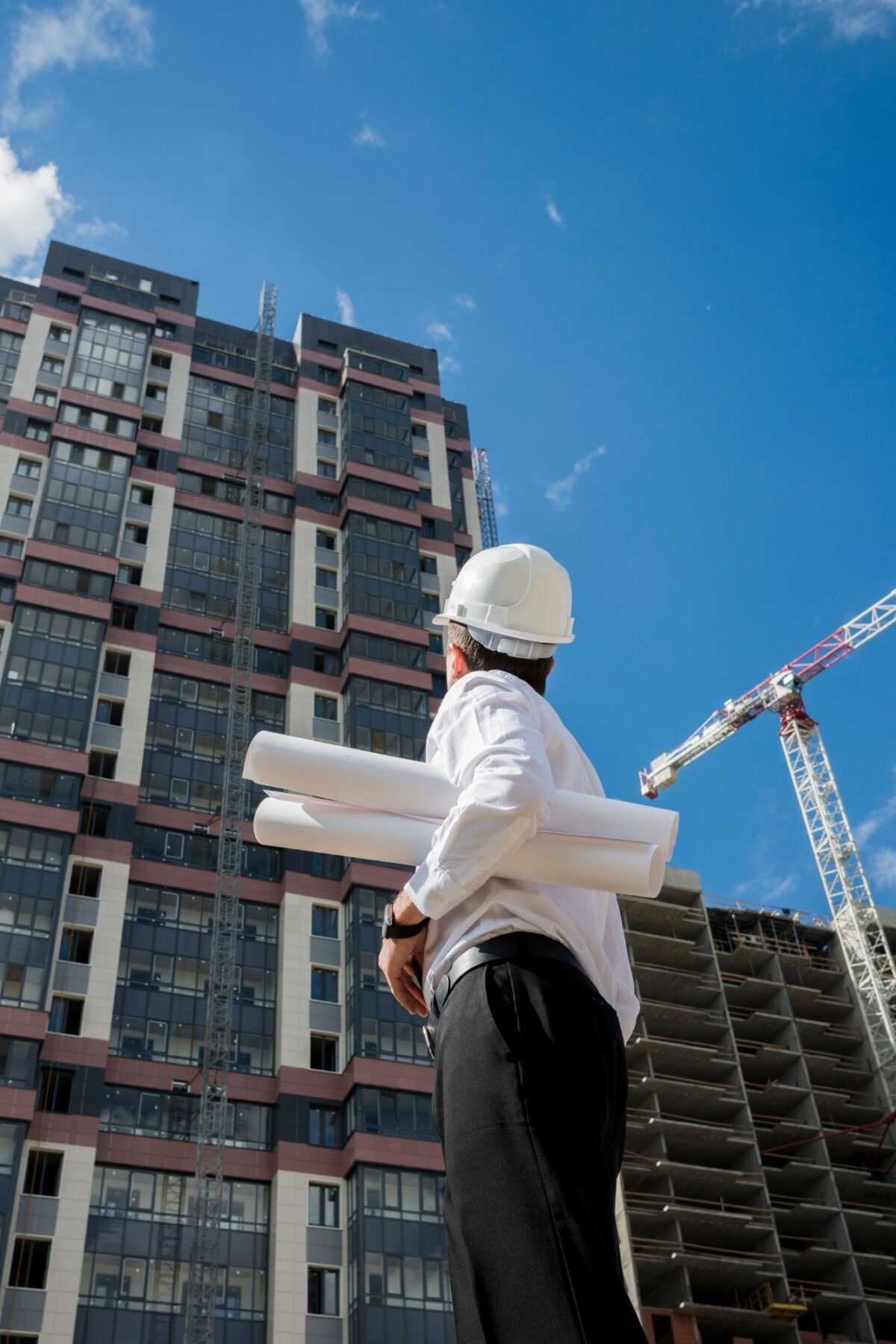 Profissional de construção com capacete branco de costas, segurando plantas arquitetônicas e observando um edifício em construção com guindastes ao fundo.