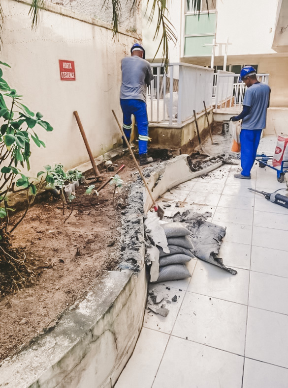 Dois operários com capacetes azuis removendo revestimento de jardineira em área interna do condomínio L’Essence Laranjeiras.