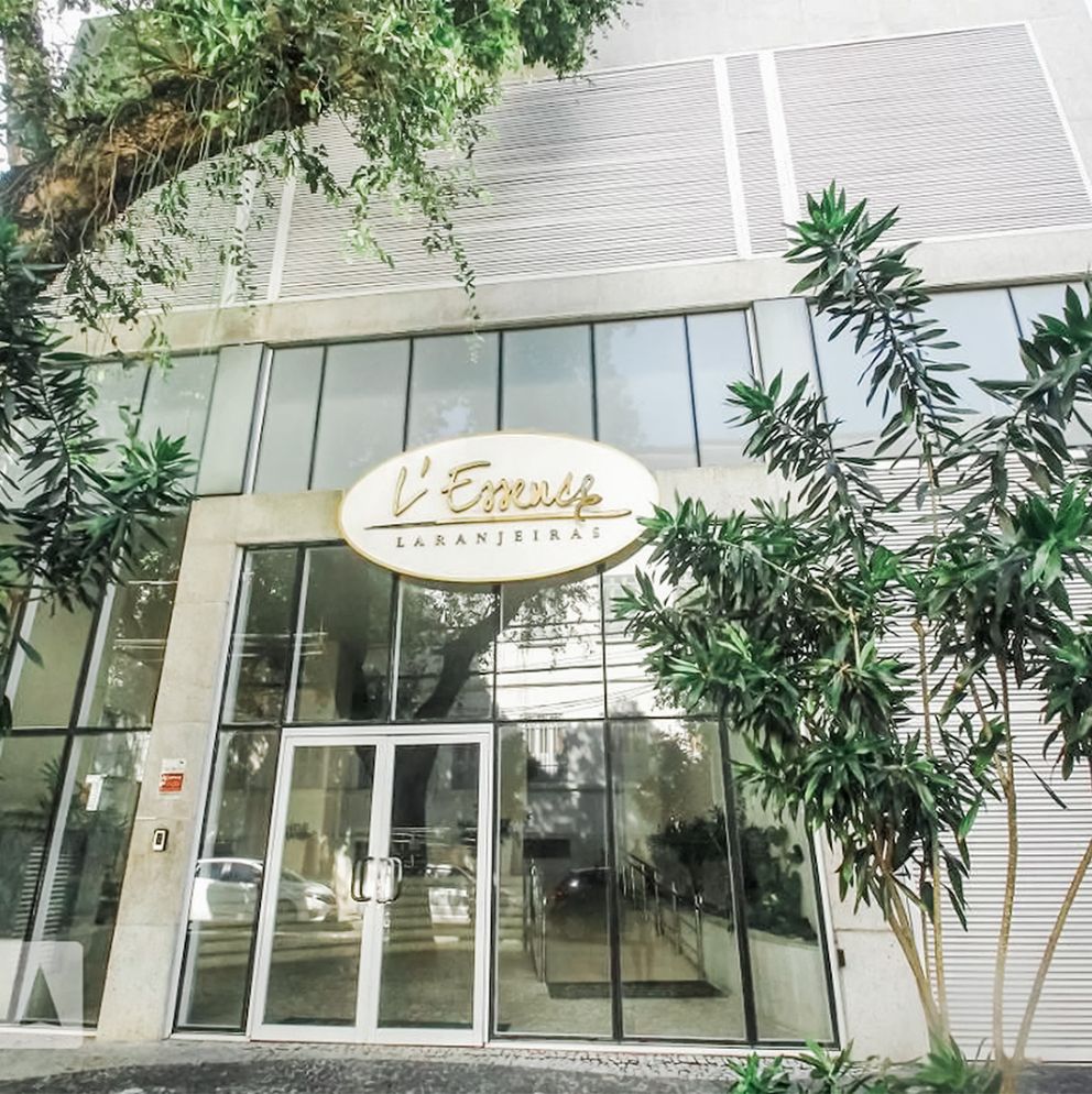 Fachada do condomínio L’Essence Laranjeiras com porta de vidro e letreiro dourado, rodeado por vegetação.