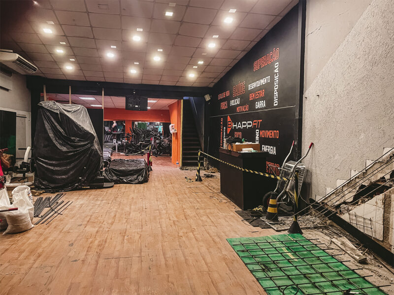 Área interna da academia antes da reforma, com piso amadeirado, equipamentos cobertos, escada lateral e estrutura em preparação para obras.