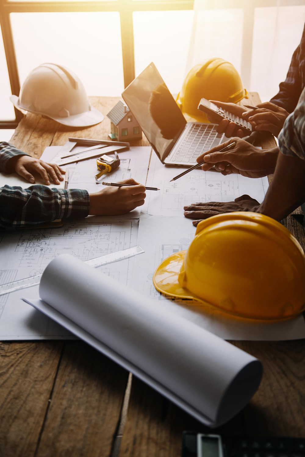 Equipe de profissionais analisando plantas e projetos de obra sobre mesa com capacetes, notebook, trena e documentos técnicos.
